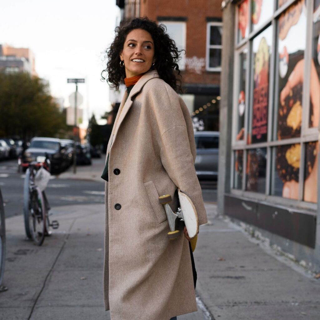 Jeune femme souriante en manteau beige, planche à roulettes sous le bras, sur un trottoir de quartier