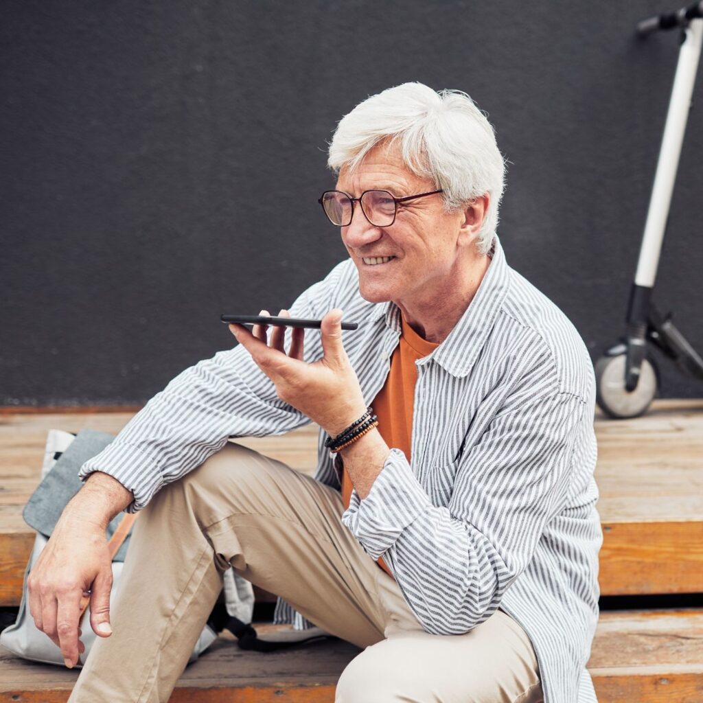 Homme aux cheveux blancs souriant, téléphone à la main, assis sur des marches en bois près d’une trottinette